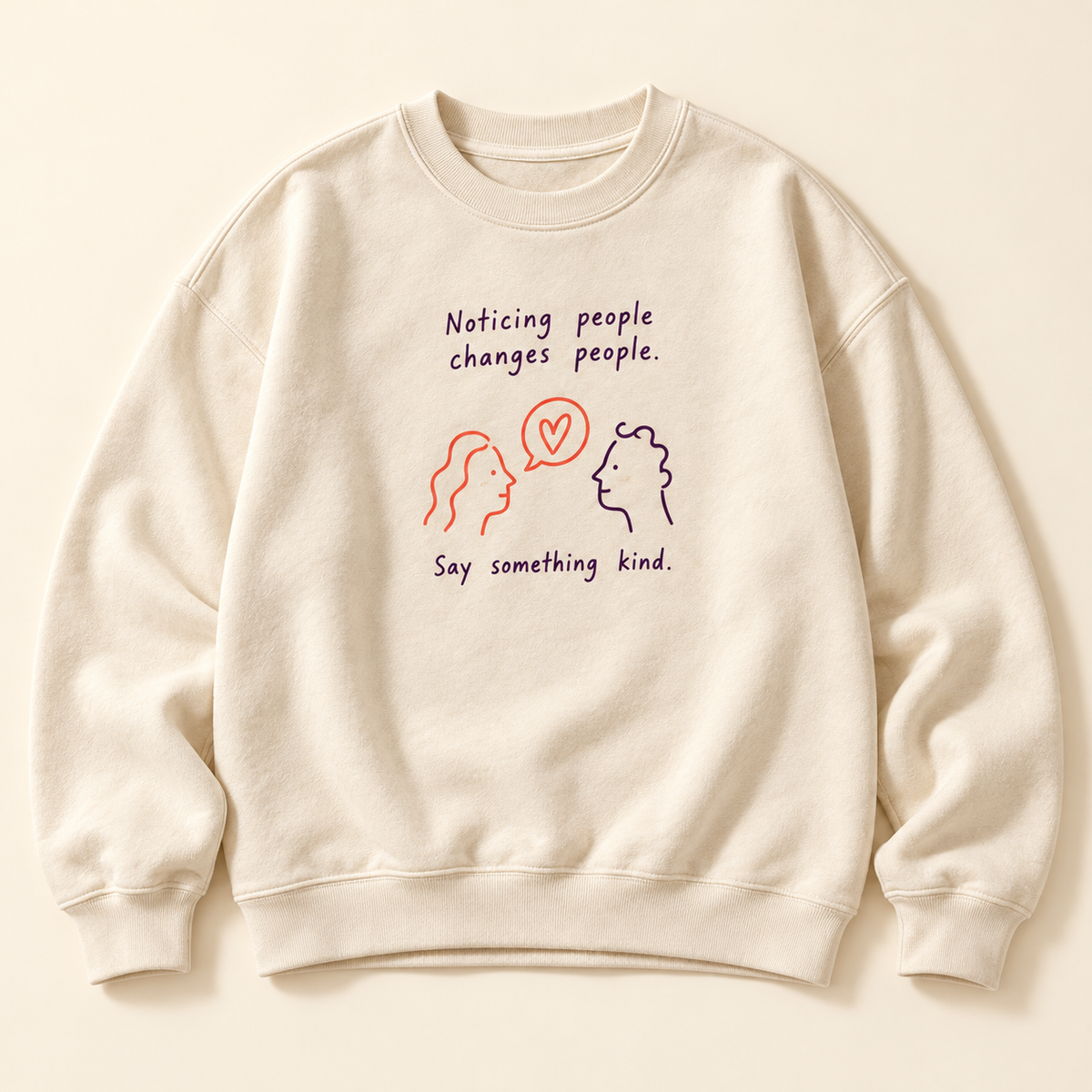 Cream crewneck with Noticing people changes people lettering, two faces and a heart speech bubble, and Say something kind beneath