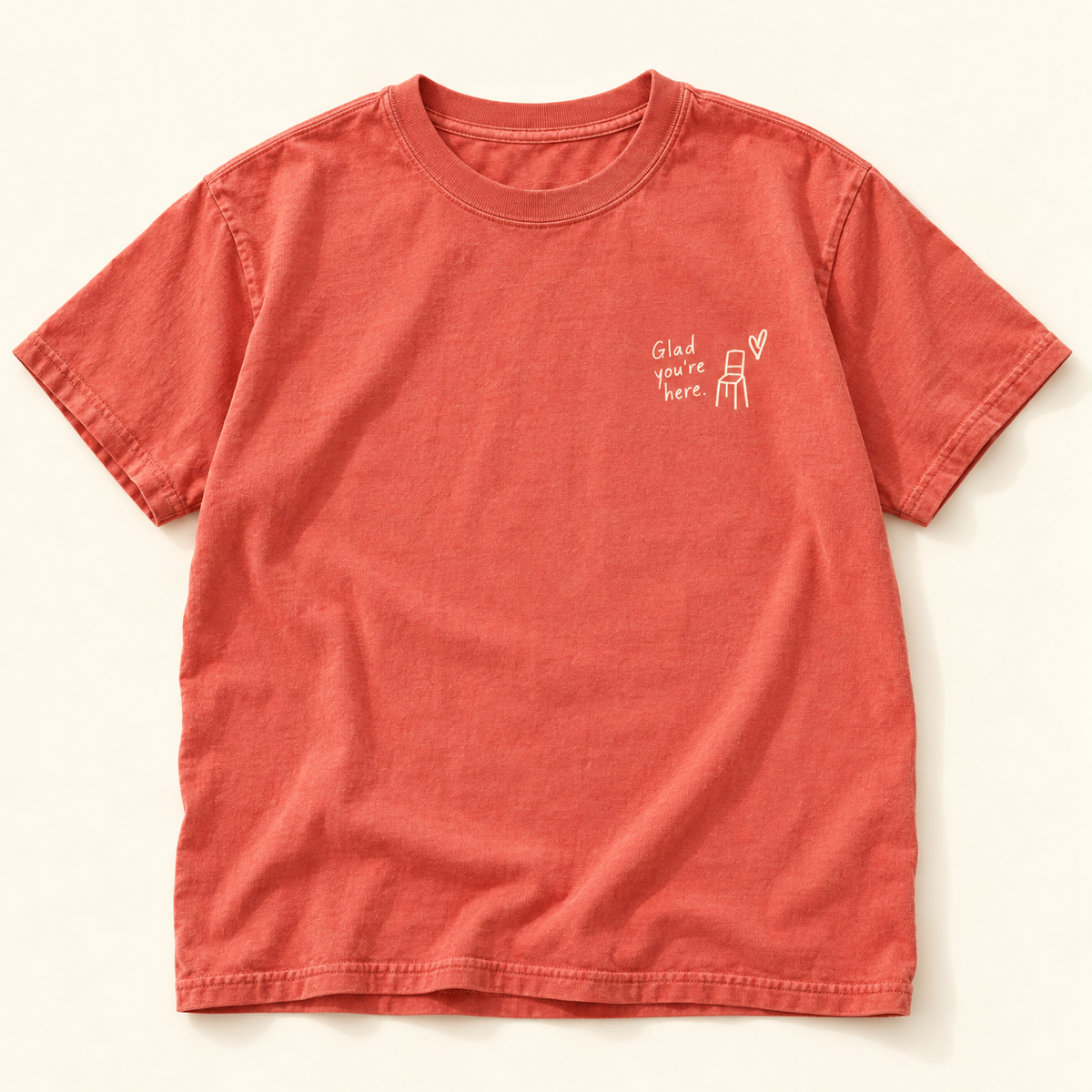 Coral tee with small cream Glad you’re here lettering, chair illustration, and heart on the chest